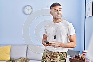 Young hispanic man army soldier using smartphone at home