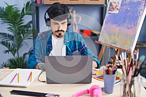 Young hispanic man artist using laptop and headphones at art studio
