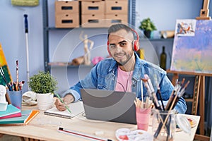 Young hispanic man artist using laptop and headphones drawing on notebook at art studio