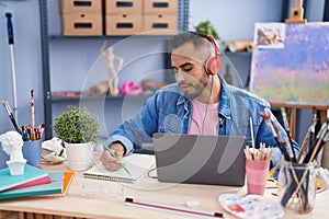 Young hispanic man artist using laptop and headphones drawing on notebook at art studio