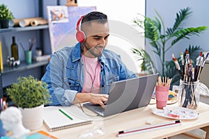 Young hispanic man artist using laptop and headphones drawing on notebook at art studio