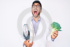 Young hispanic man as nutritionist doctor holding weighing machine and broccoli angry and mad screaming frustrated and furious,