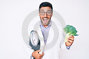 Young hispanic man as nutritionist doctor holding weighing machine and broccoli celebrating crazy and amazed for success with open