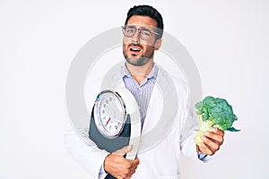 Young hispanic man as nutritionist doctor holding weighing machine and broccoli clueless and confused expression