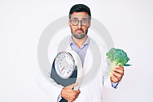 Young hispanic man as nutritionist doctor holding weighing machine and broccoli depressed and worry for distress, crying angry and