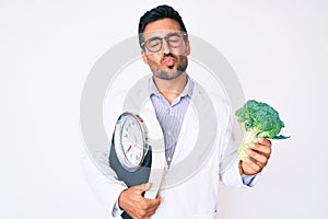 Young hispanic man as nutritionist doctor holding weighing machine and broccoli looking at the camera blowing a kiss being lovely