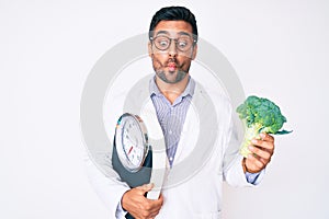 Young hispanic man as nutritionist doctor holding weighing machine and broccoli making fish face with mouth and squinting eyes,