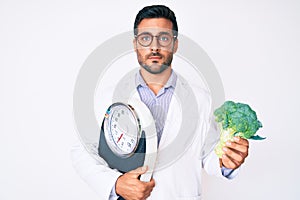 Young hispanic man as nutritionist doctor holding weighing machine and broccoli relaxed with serious expression on face