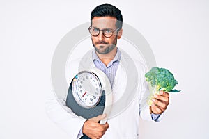 Young hispanic man as nutritionist doctor holding weighing machine and broccoli skeptic and nervous, frowning upset because of