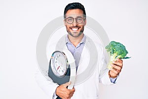 Young hispanic man as nutritionist doctor holding weighing machine and broccoli smiling with a happy and cool smile on face