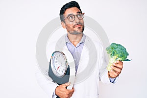 Young hispanic man as nutritionist doctor holding weighing machine and broccoli smiling looking to the side and staring away