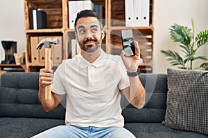 Young hispanic man with beard holding hammer and broken smartphone showing cracked screen smiling looking to the side and staring
