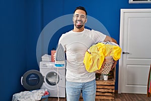 Young hispanic man with beard holding iron and clothes at home sticking tongue out happy with funny expression