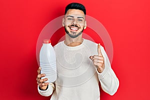 Young hispanic man with beard holding liter bottle of milk smiling with an idea or question pointing finger with happy face,