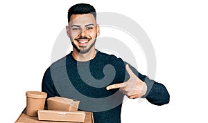 Young hispanic man with beard holding take away food smiling happy pointing with hand and finger