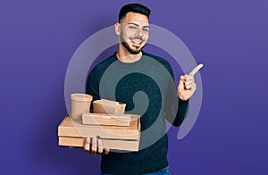 Young hispanic man with beard holding take away food smiling happy pointing with hand and finger to the side