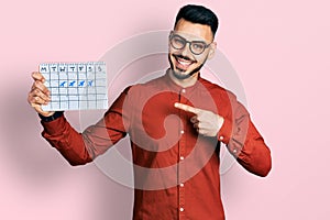 Young hispanic man with beard holding travel calendar smiling happy pointing with hand and finger