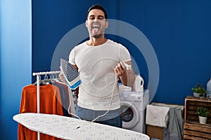 Young hispanic man with beard ironing clothes at home sticking tongue out happy with funny expression