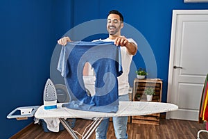 Young hispanic man with beard ironing holding burned iron shirt at laundry room smiling and laughing hard out loud because funny