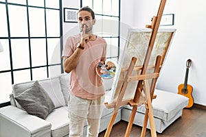 Young hispanic man with beard painting on canvas at home asking to be quiet with finger on lips