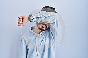 Young hispanic man with beard standing over blue background covering eyes with arm, looking serious and sad