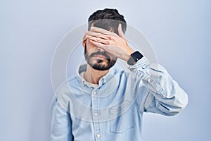 Young hispanic man with beard standing over blue background covering eyes with hand, looking serious and sad