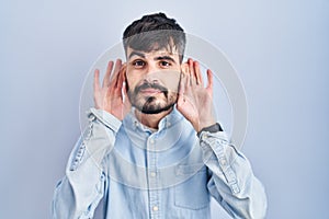 Young hispanic man with beard standing over blue background trying to hear both hands on ear gesture, curious for gossip