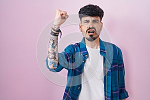 Young hispanic man with beard standing over pink background angry and mad raising fist frustrated and furious while shouting with