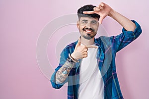 Young hispanic man with beard standing over pink background smiling making frame with hands and fingers with happy face