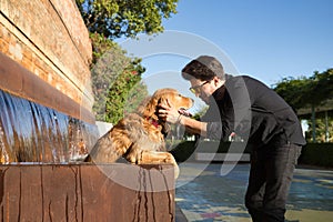 Young Hispanic man with a beard, sunglasses and black shirt, petting his wet dog that has gotten into a fountain because of the