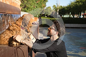 Young Hispanic man with a beard, sunglasses and black shirt, petting his wet dog that has gotten into a fountain because of the