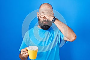 Young hispanic man with beard and tattoos drinking a cup of coffee covering eyes with hand, looking serious and sad