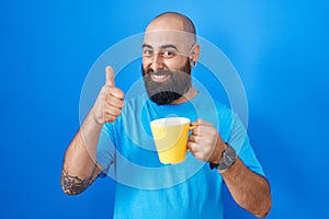 Young hispanic man with beard and tattoos drinking a cup of coffee doing happy thumbs up gesture with hand