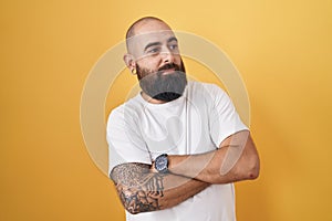 Young hispanic man with beard and tattoos standing over yellow background looking to the side with arms crossed convinced and