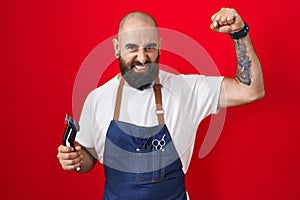 Young hispanic man with beard and tattoos wearing barber apron holding razor strong person showing arm muscle, confident and proud