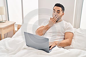 Young hispanic man with beard using computer laptop on the bed covering mouth with hand, shocked and afraid for mistake
