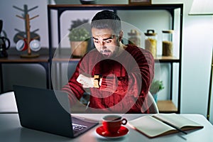 Young hispanic man with beard using computer laptop at night at home looking at the watch time worried, afraid of getting late