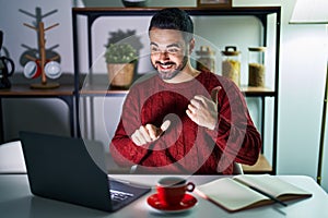 Young hispanic man with beard using computer laptop at night at home pointing to the back behind with hand and thumbs up, smiling