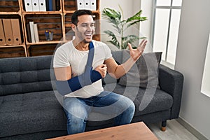 Young hispanic man with beard wearing arm on sling sitting at therapy consult celebrating victory with happy smile and winner