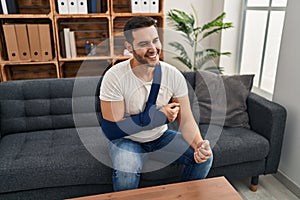 Young hispanic man with beard wearing arm on sling sitting at therapy consult looking positive and happy standing and smiling with