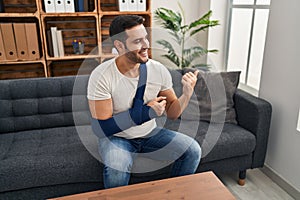 Young hispanic man with beard wearing arm on sling sitting at therapy consult pointing thumb up to the side smiling happy with