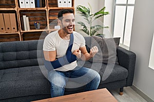 Young hispanic man with beard wearing arm on sling sitting at therapy consult screaming proud, celebrating victory and success
