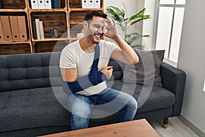 Young hispanic man with beard wearing arm on sling sitting at therapy consult smiling happy doing ok sign with hand on eye looking