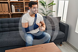 Young hispanic man with beard wearing arm on sling sitting at therapy consult smiling happy pointing with hand and finger