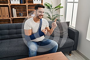 Young hispanic man with beard wearing arm on sling sitting at therapy consult smiling happy pointing with hand and finger to the