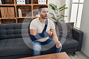 Young hispanic man with beard wearing arm on sling sitting at therapy consult thinking attitude and sober expression looking self