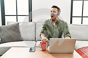 Young hispanic man with beard wearing call center agent headset working from home angry and mad screaming frustrated and furious,