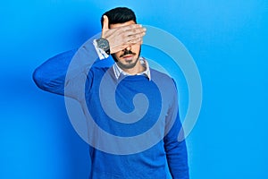 Young hispanic man with beard wearing casual blue sweater covering eyes with hand, looking serious and sad