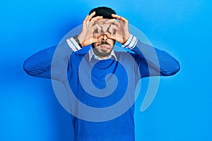 Young hispanic man with beard wearing casual blue sweater doing ok gesture like binoculars sticking tongue out, eyes looking