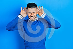 Young hispanic man with beard wearing casual blue sweater trying to hear both hands on ear gesture, curious for gossip